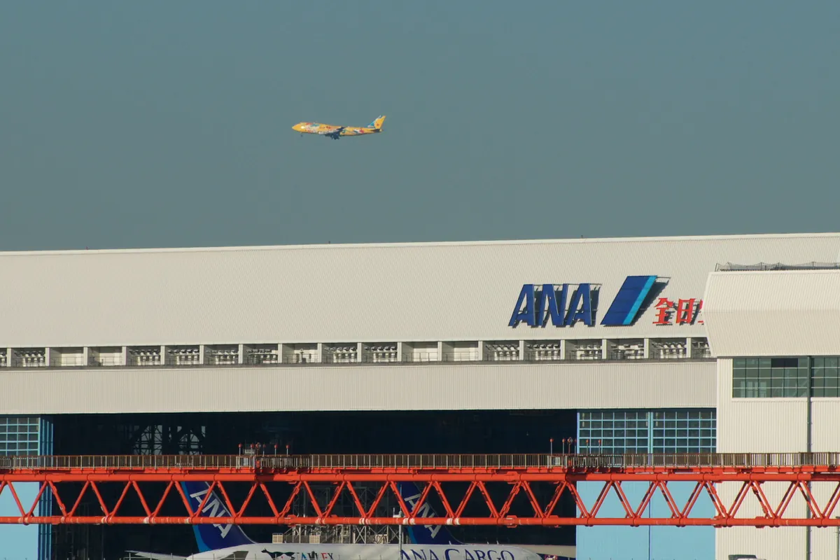ANAの整備工場の建物に「ANA」のロゴが大きく掲げられ、上空を特別塗装の旅客機が飛行している写真