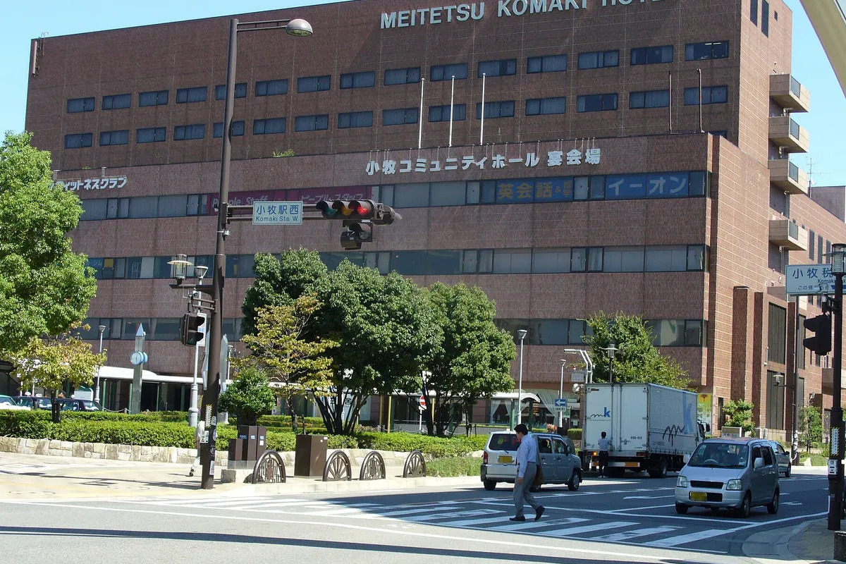 小牧駅西口の交差点で、名鉄小牧ホテルの建物と横断歩道、信号機、トラックや乗用車が並ぶ様子の写真