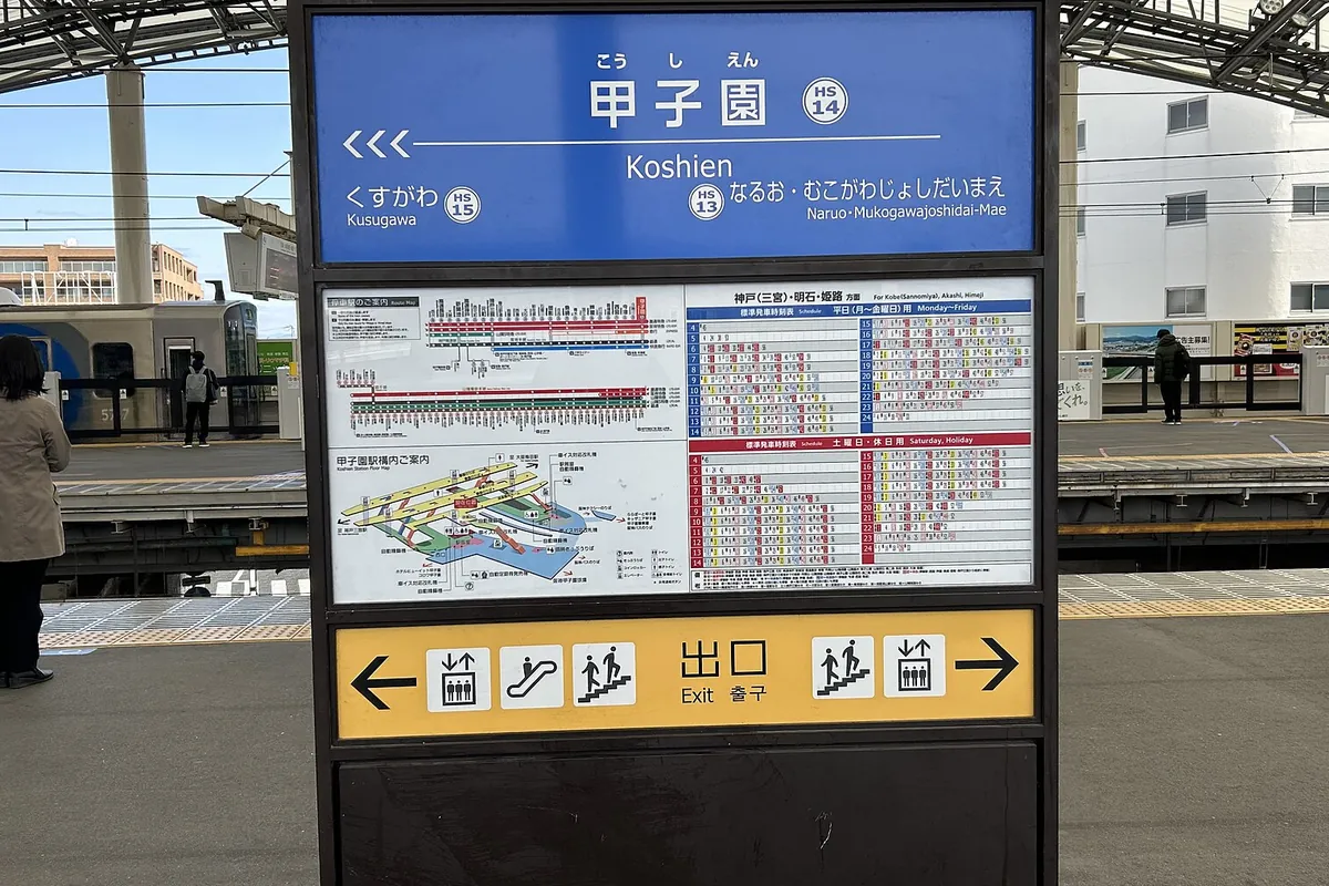 阪神甲子園駅のホームにある青い駅名標と時刻表の案内板、奥に停車中の電車が写る写真