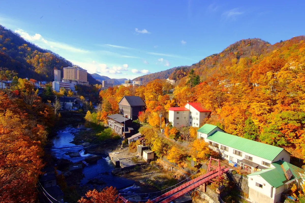 北海道の定山渓温泉で、渓谷沿いに赤い吊り橋と旅館の建物があり、山一面が紅葉している様子の写真