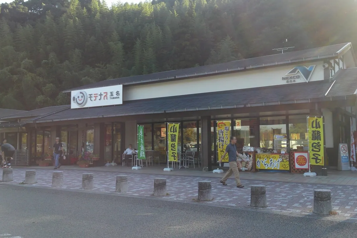 九州自動車道・玉名パーキングエリアの店舗外観で、「モテナス玉名」の看板と山鹿鶏の幟が並ぶ様子の写真
