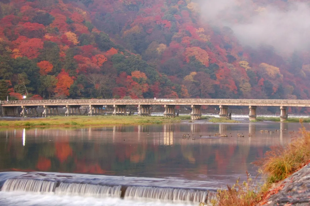渡月橋と周辺の山が紅葉で赤や橙に色づき、川面にその様子が映り込む景色の写真