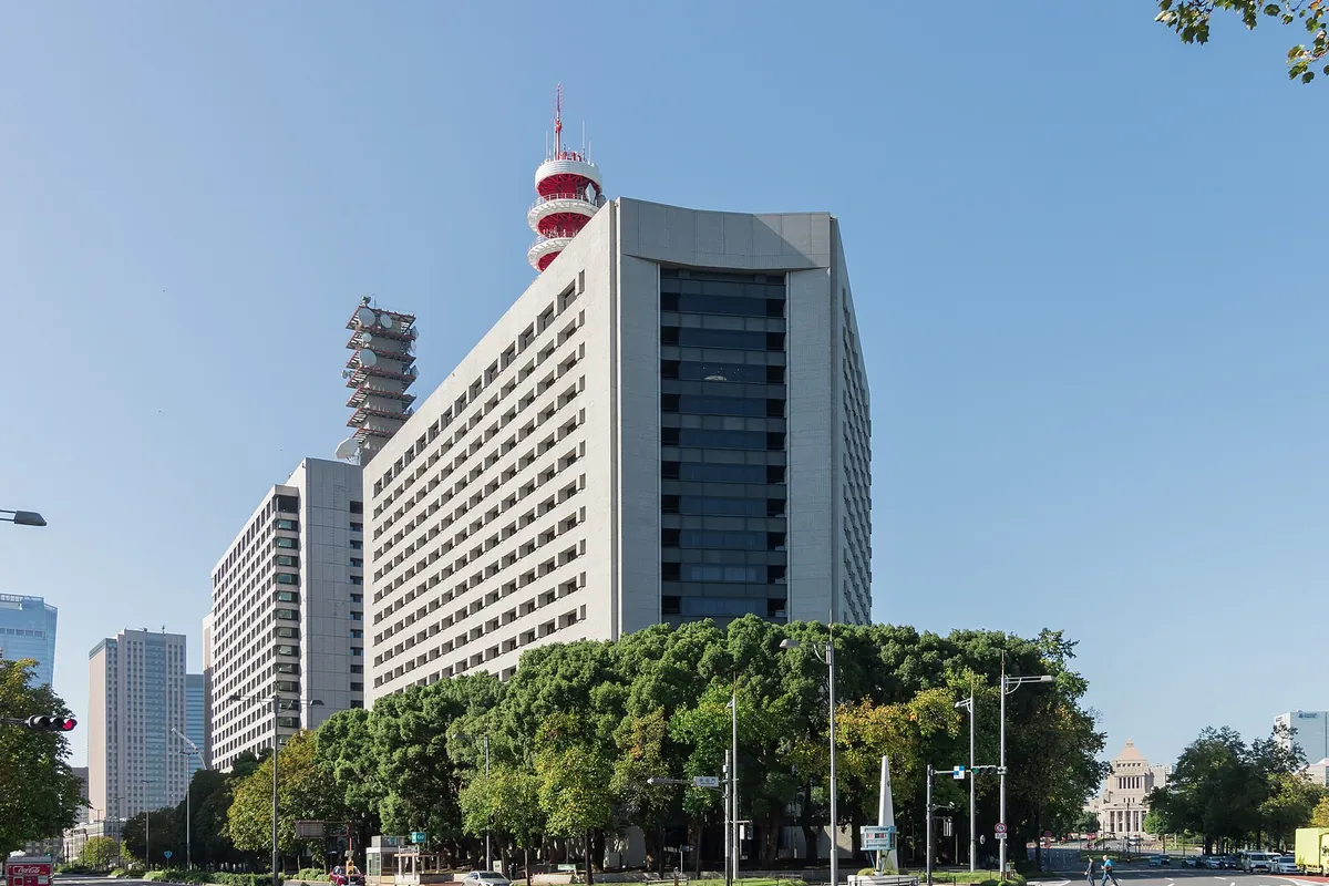 赤白の電波塔が屋上に立つ高層庁舎ビルの外観と、手前の街路樹や国会議事堂らしき建物が見える様子の写真