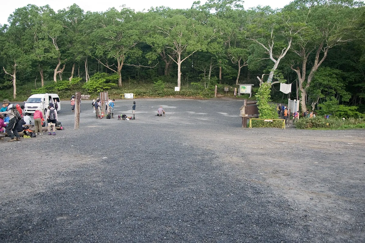 森に囲まれた鳩待峠の駐車場に登山客が数人集まり、荷物を整えたり休憩したりしている様子の写真