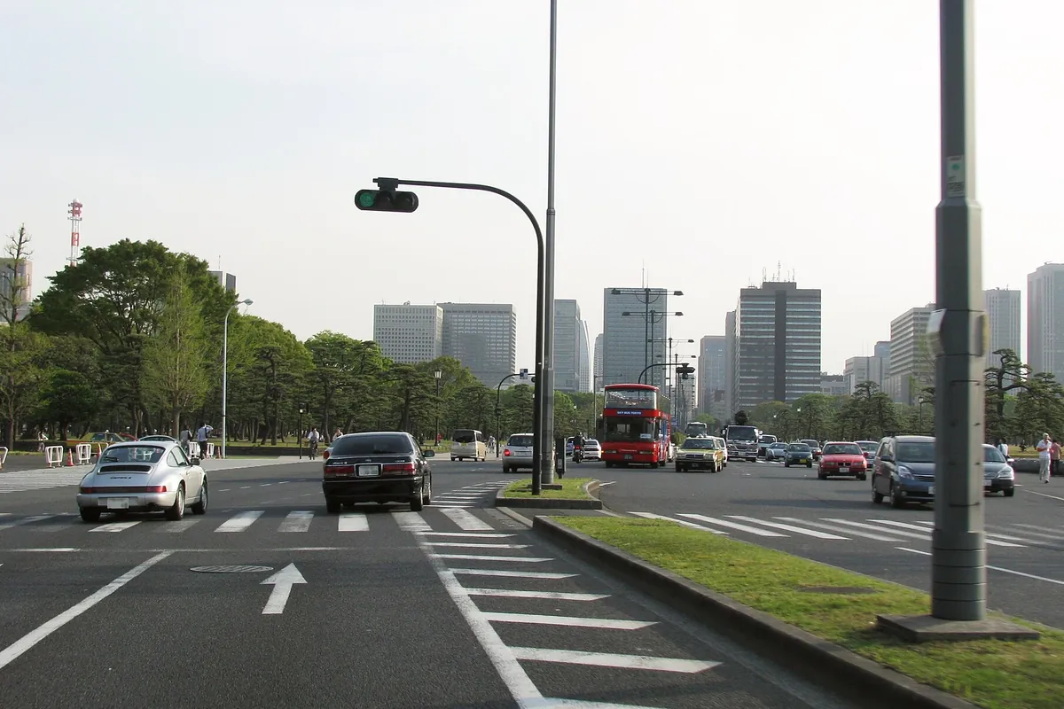 東京の皇居周辺の通りで、赤い二階建てバスや乗用車が走り、奥に高層ビル群が見える様子の写真