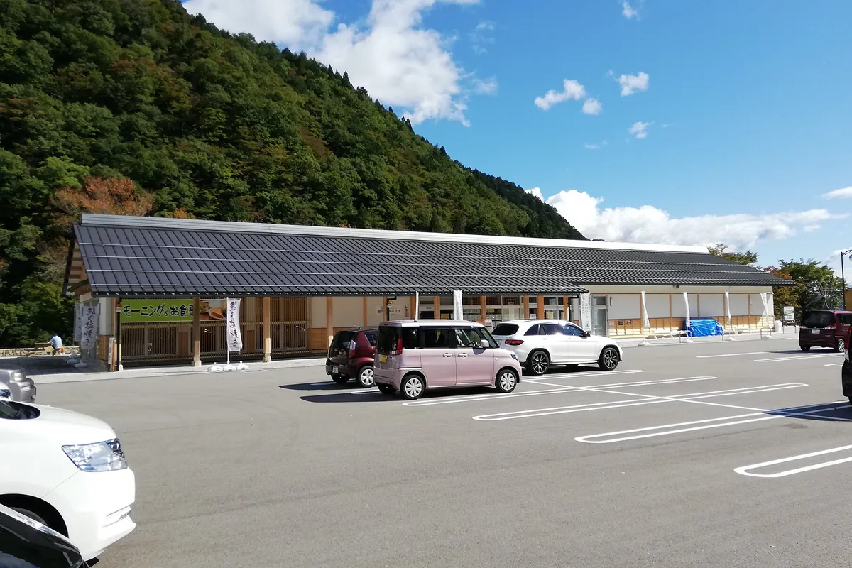 山のふもとに建つ道の駅の木造の建物の前に、乗用車が数台停まった駐車場が広がる写真