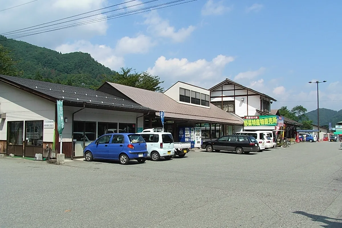 山を背にした道の駅の建物が並び、「野菜特産物直売所」の看板が見え、前の駐車場に車が停まっている写真