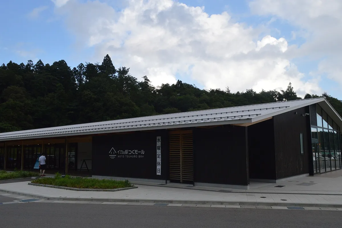 黒い外壁の建物に「イカの駅つくモール NOTO TSUKUMO BAY」と書かれた看板が掲げられ、背後に木々の茂る丘が広がる写真