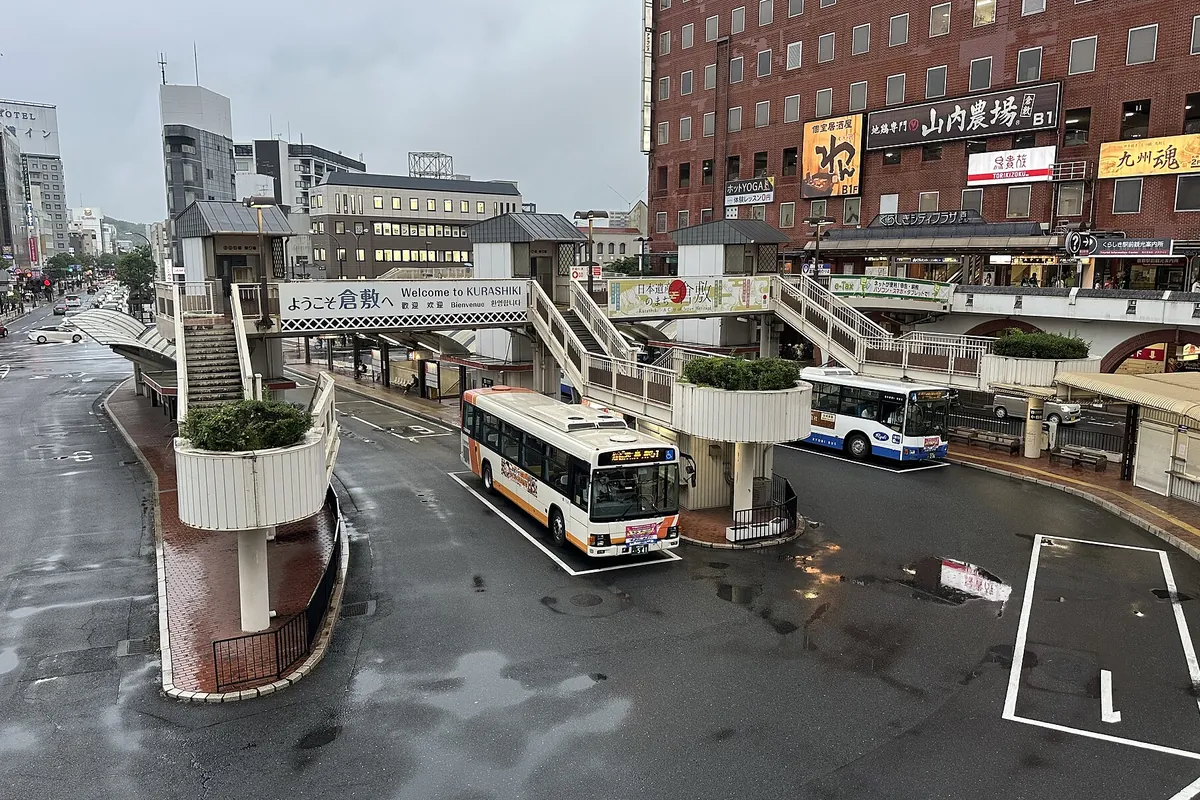 倉敷駅のバスターミナルで、雨の中「ようこそ倉敷へ」の看板下に路線バスが2台停車している様子の写真