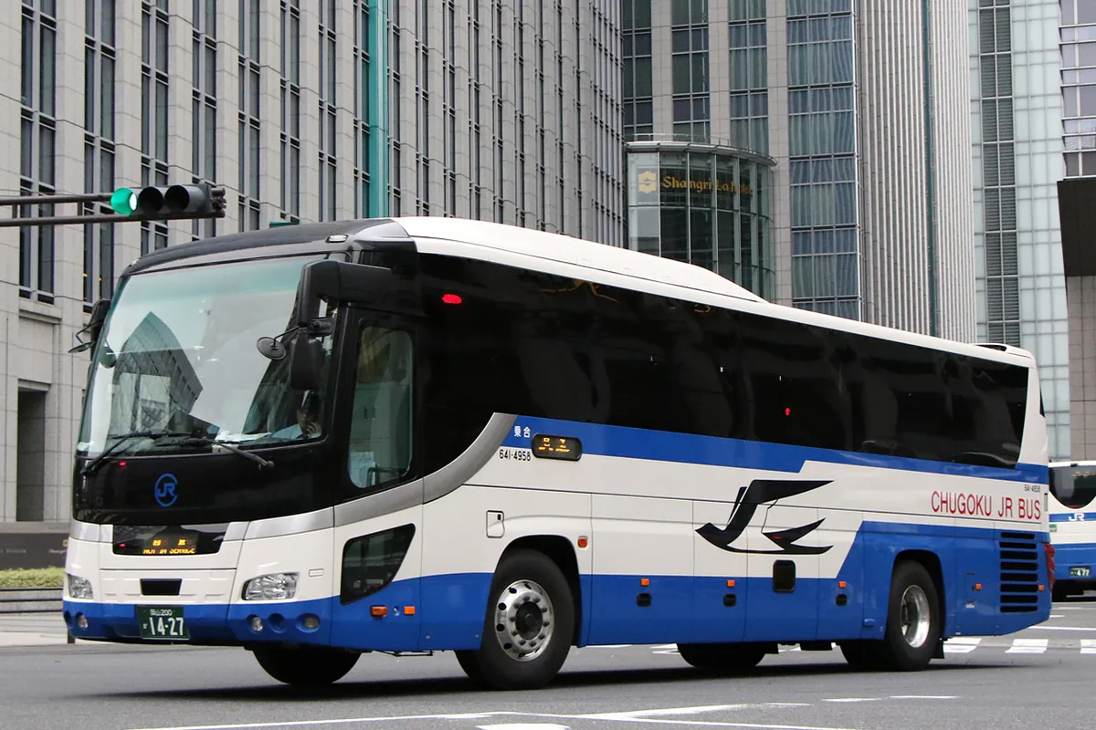 「CHUGOKU JR BUS」と書かれた青と白の夜行高速バスが、ビル街の交差点を走行している様子の写真