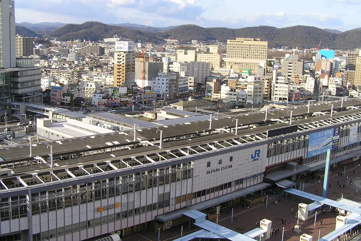 「岡山駅 OKAYAMA STATION」の駅舎と線路を俯瞰し、周囲の市街地やJR岡山駅前広場が広がる様子の写真