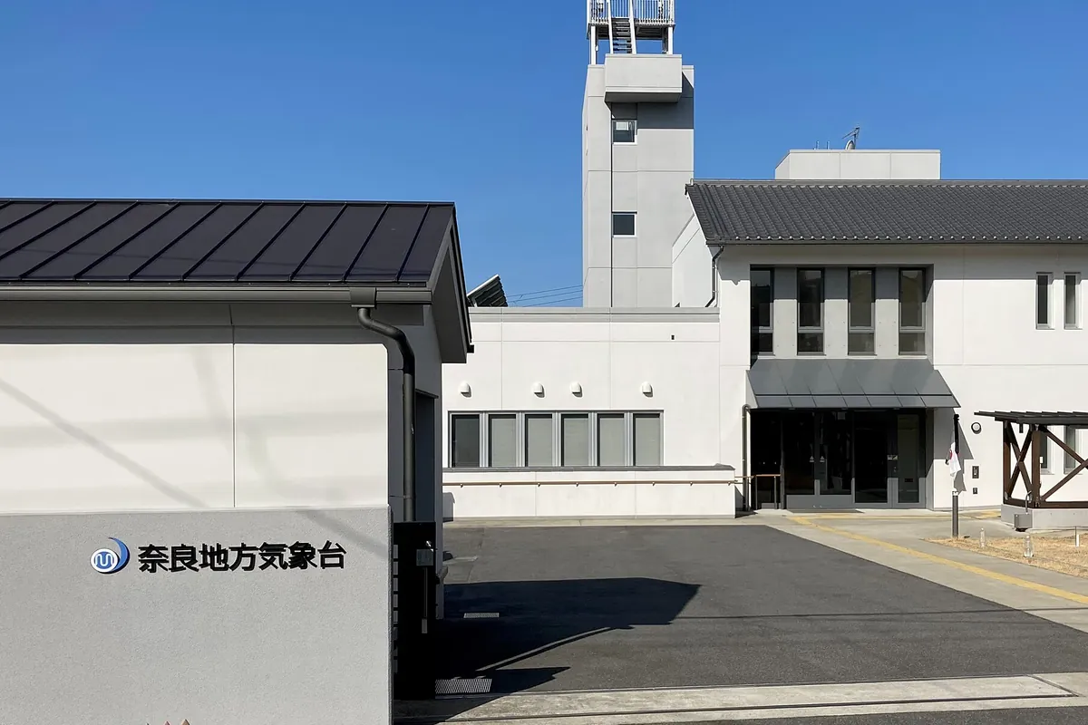白い壁の建物に「奈良地方気象台」と書かれた看板があり、奥に展望台のような塔がそびえる庁舎の写真