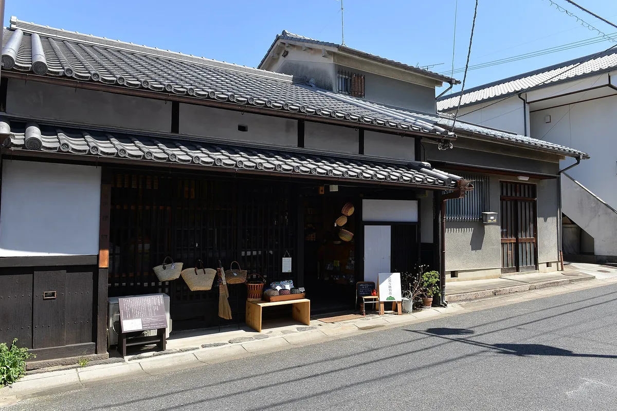 瓦屋根の古い町家が並ぶ通りに、かごや小物を並べた商店の軒先が面する街並みの写真