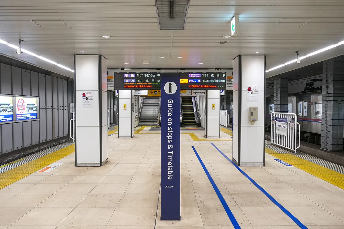 京成電鉄の駅ホームで、案内柱越しに「成田」「ちはら台」行きの発車案内と停車中の列車が見える写真