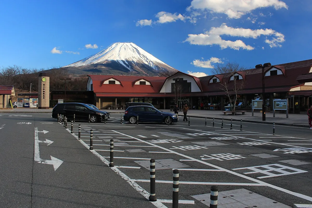 赤い屋根の建物の背後に雪をかぶった富士山がそびえ、手前に駐車場と車が並ぶ道の駅の写真
