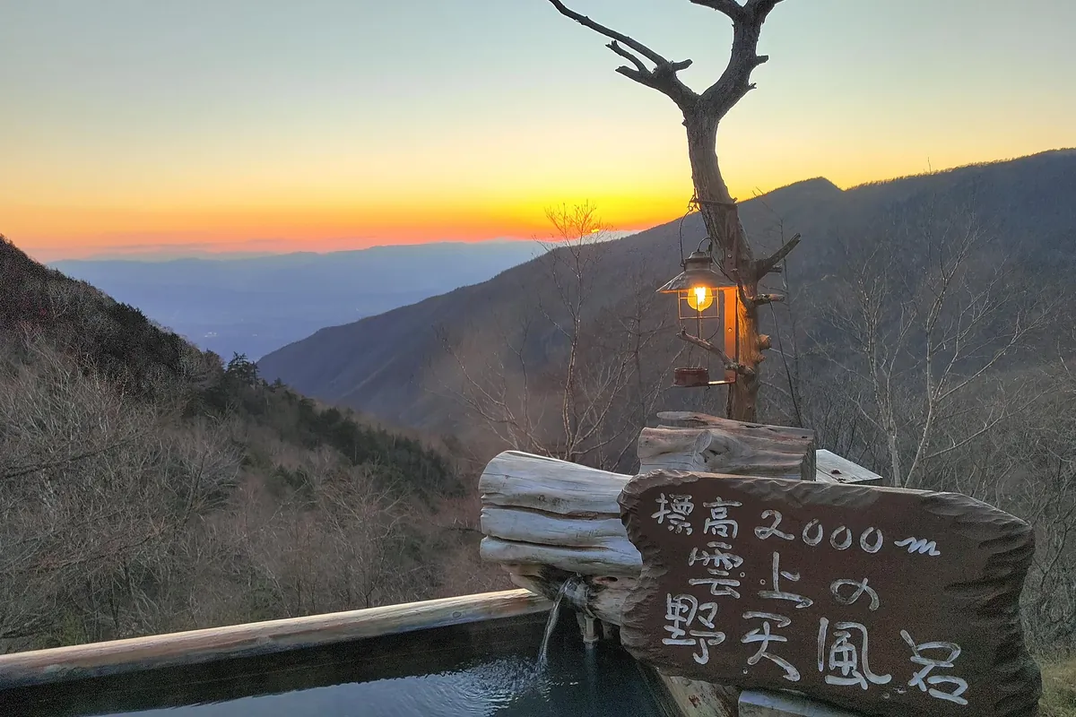 山並みに夕日が沈む景色を望む、標高2000mの野天風呂と書かれた木の湯船の写真