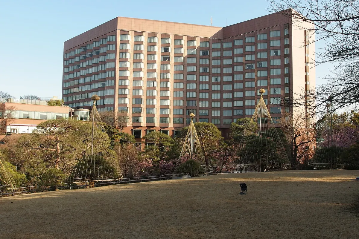 日本庭園に雪吊りが並ぶ手前の芝生広場越しに、高層ホテルの建物外観を見た写真