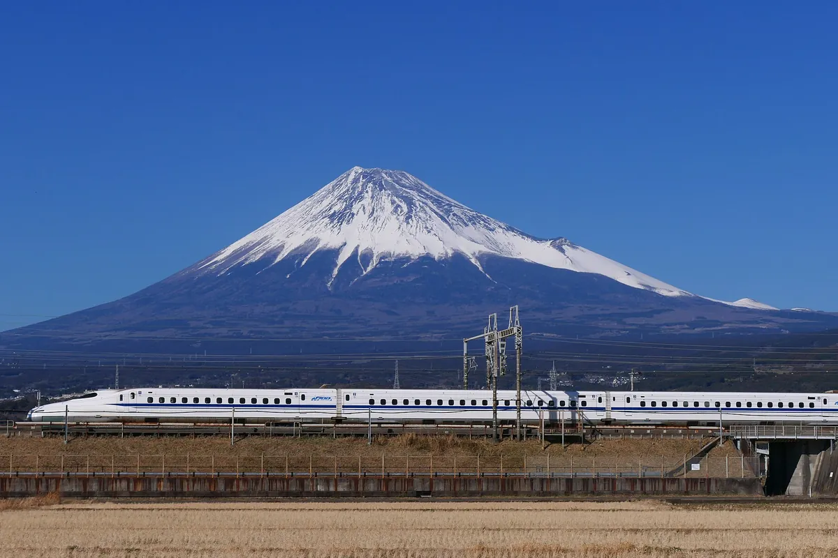 雪をかぶった富士山を背景に、東海道新幹線の白とブルーの車両が高架線を走行している写真