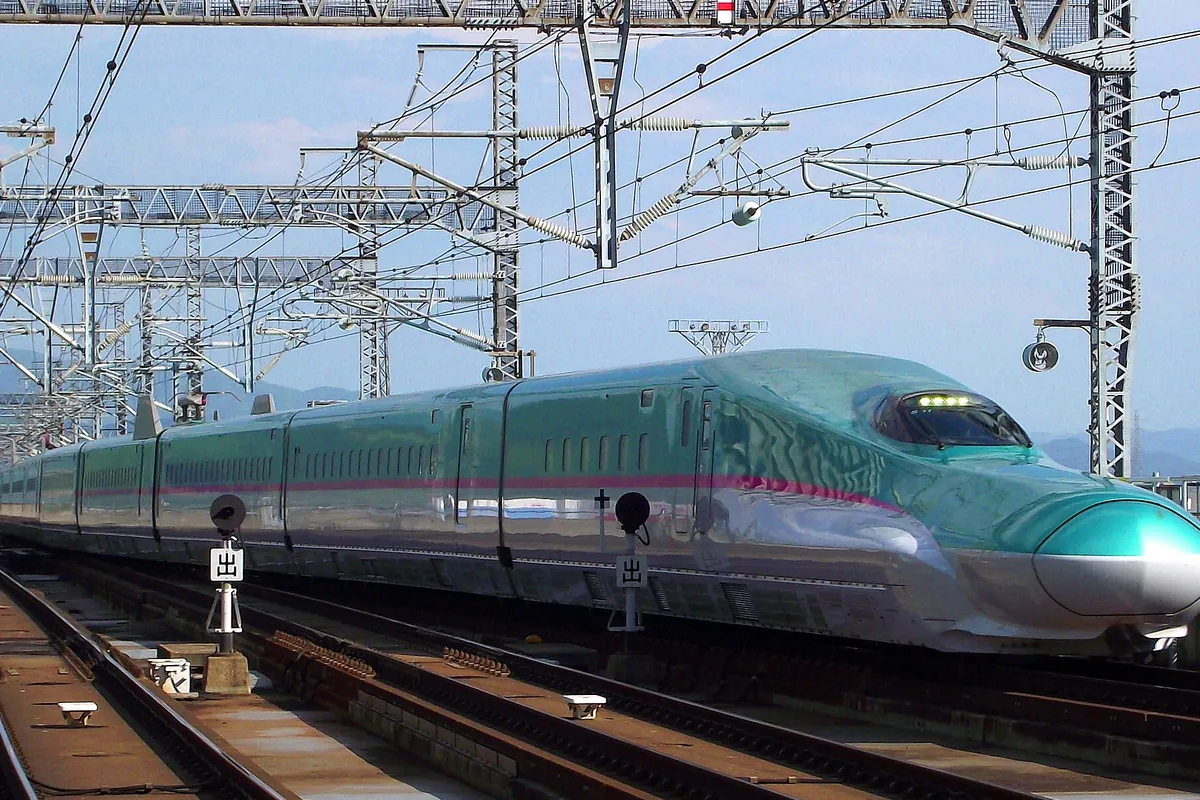 高架の架線柱が並ぶ線路脇を走る緑色とピンクのラインが入った新幹線車両の写真