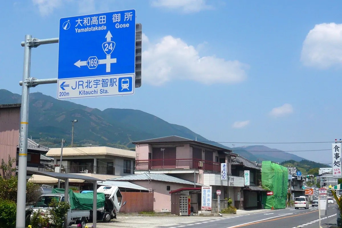 国道24号・国道169号方面を示す青い案内標識と住宅が並ぶ道路沿いの街並みの写真