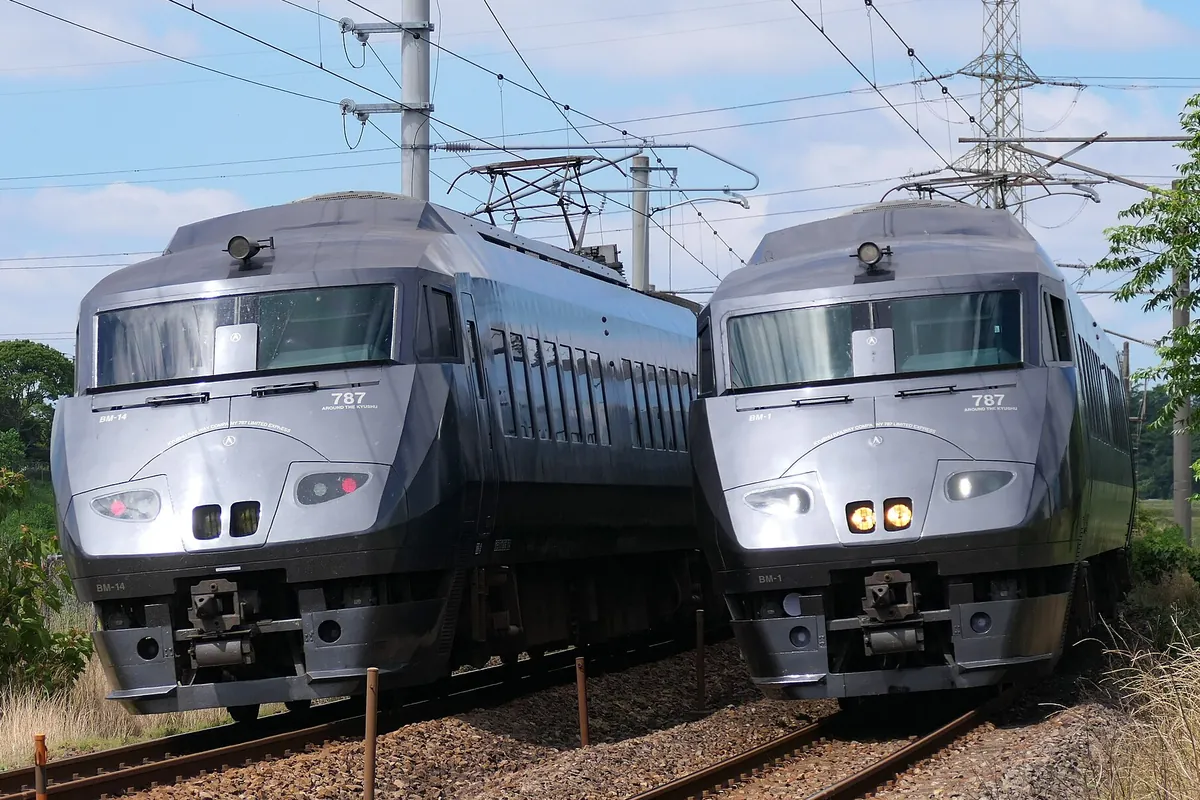 「787系」「AROUND THE KYUSHU」と表示された銀色の特急車両が線路上に2編成並んで停車している写真