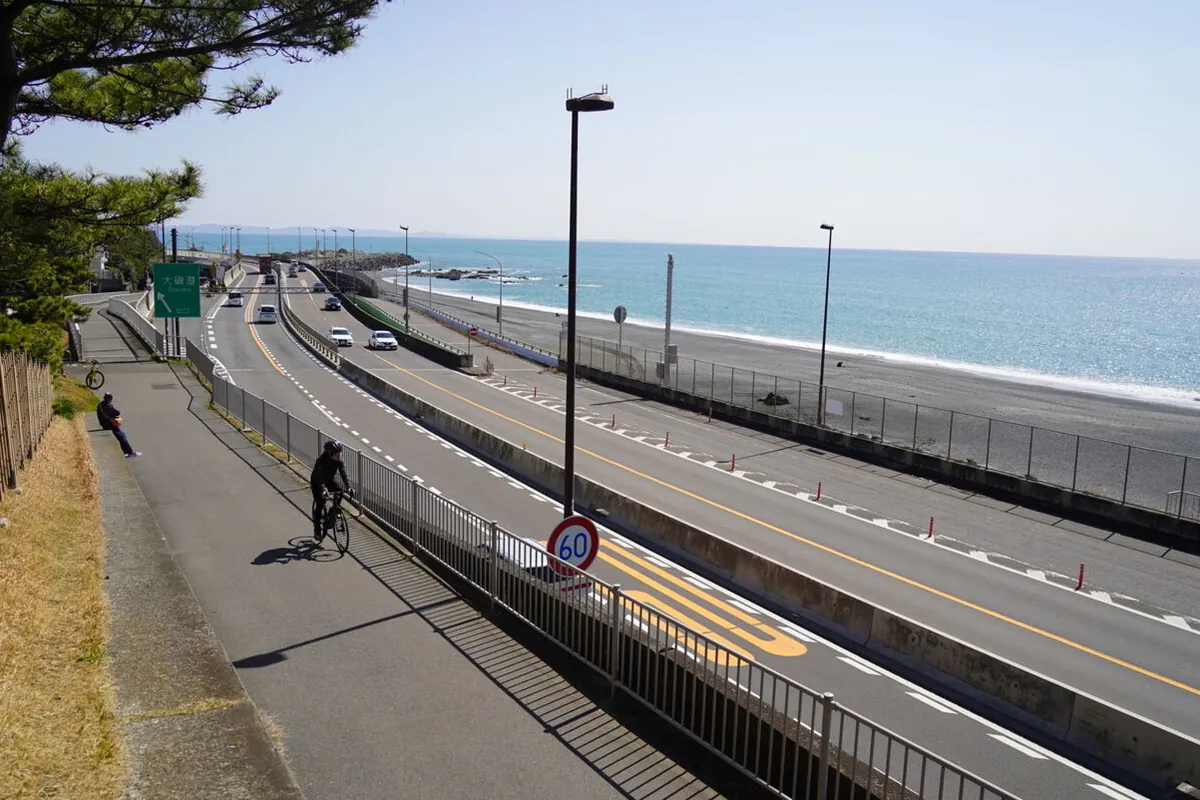 海沿いの片側二車線の道路が緩やかに右へカーブし、フェンスの向こうに砂浜と海が広がる景観の写真
