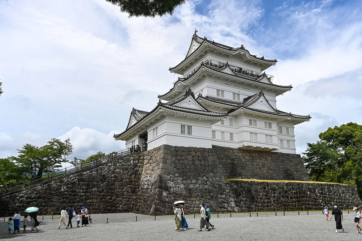 石垣の上に建つ白壁の三層の天守閣を見上げる構図で、手前の広場を観光客が歩いている様子の写真