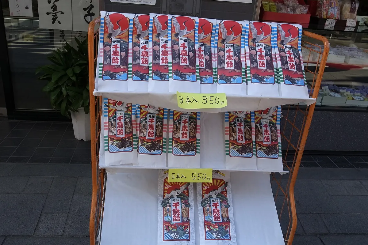 和菓子店の店頭に並ぶ「元祖千歳飴」と書かれた包装の千歳飴と、価格を示す値札が写る写真