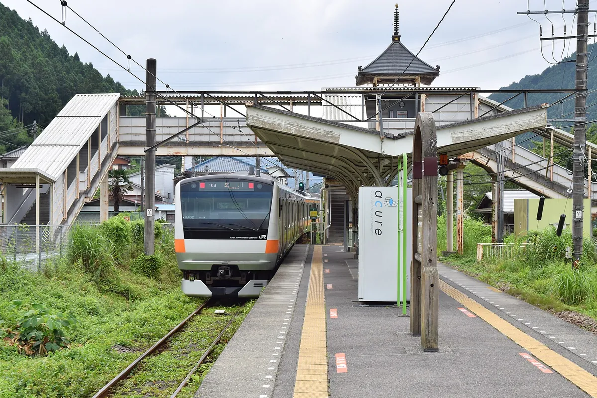 山に囲まれた奥多摩駅のホームに、オレンジ色の帯が入った青梅線の電車が停車している写真