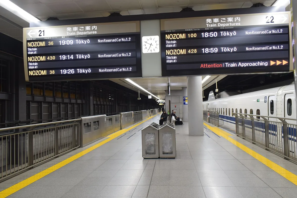 新幹線の駅のホームで、発車案内の電光掲示板とNOZOMIの到着を待つ乗客、停車中の新幹線車両が写る写真