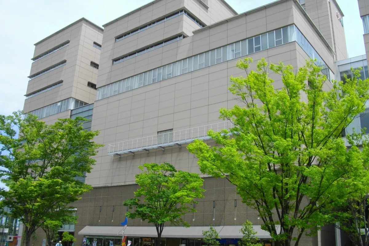 彩の国ビジュアルプラザの建物外観を、手前に新緑の木々が並ぶ歩道から見上げて撮った写真