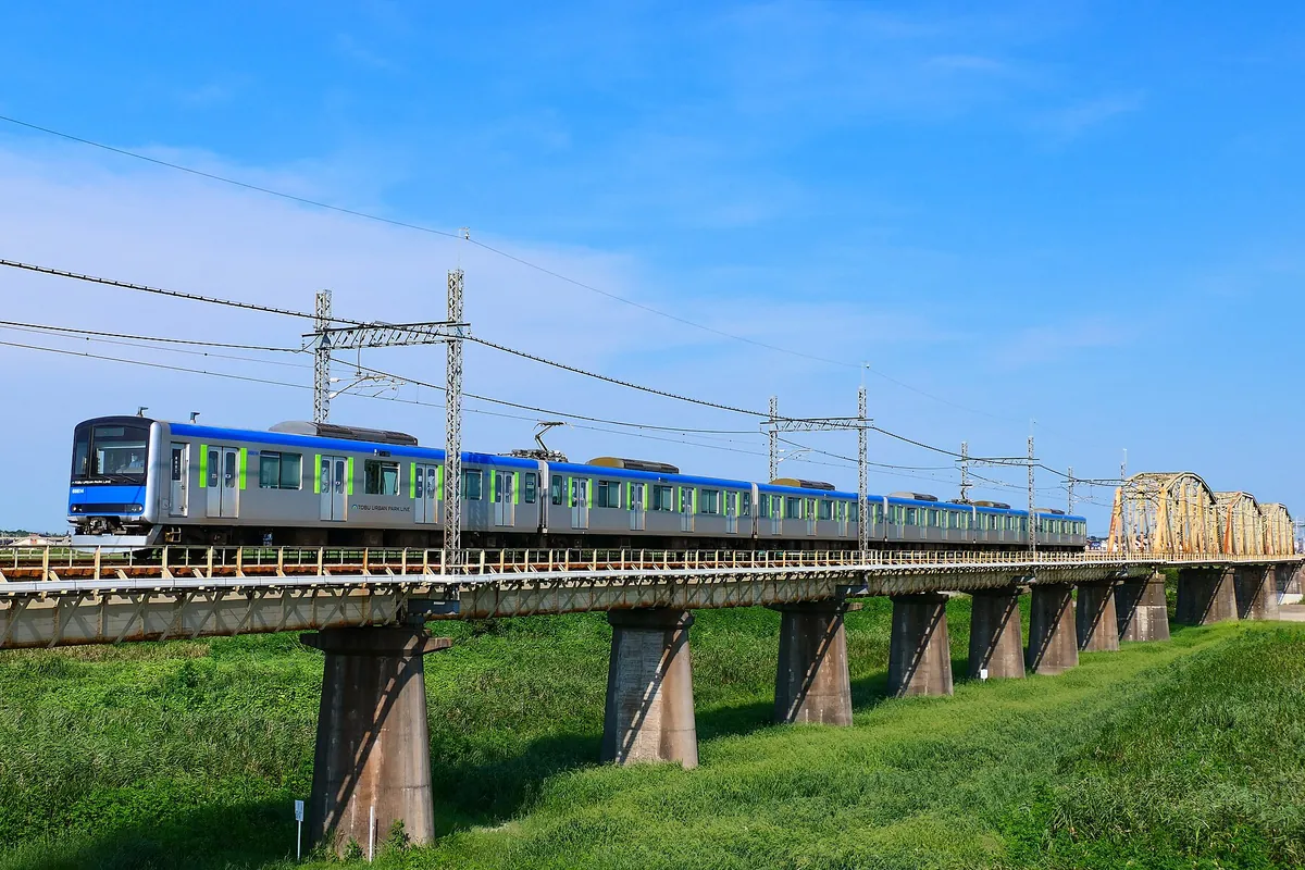 河川に架かる鉄橋を青と緑のラインの「TOBU URBAN PARK LINE」の電車が走行する様子の写真