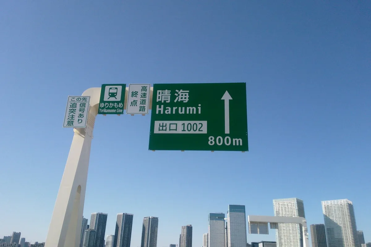 首都高速晴海線の「晴海 出口1002 800m」と書かれた案内標識がビル群の上に掲げられている写真