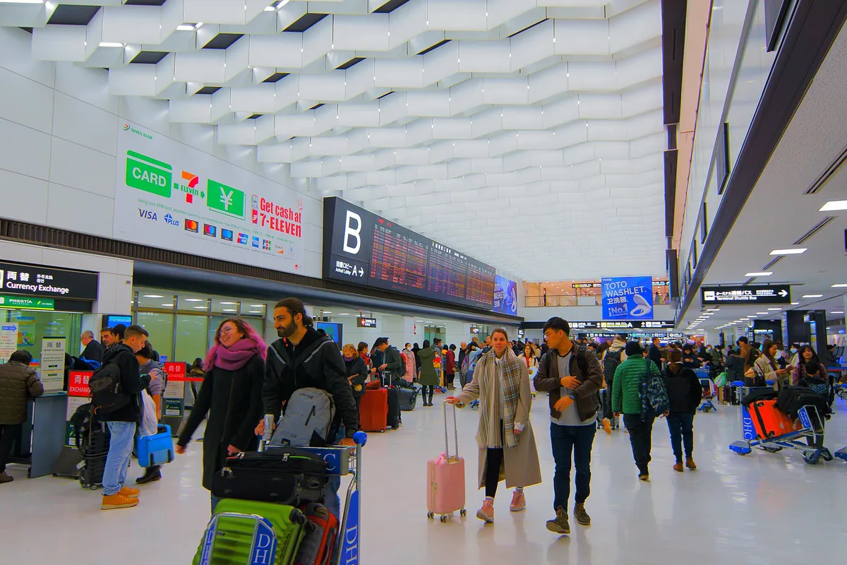 成田空港の出発ロビーで、両替カウンターやスーツケースを押す旅行客たちが行き交う様子の写真