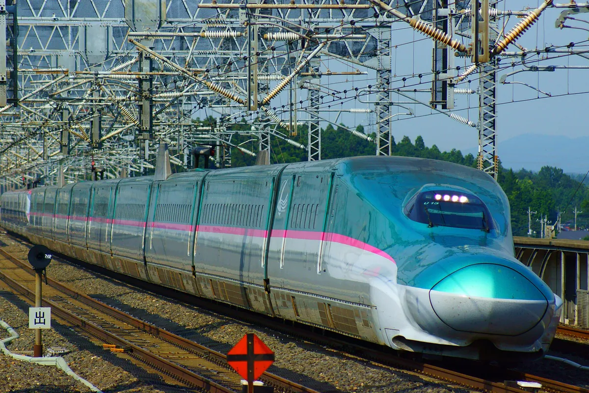 高架の架線設備が並ぶ区間を走る、ピンク色の帯が入ったJR東日本の新幹線車両の先頭部分の写真
