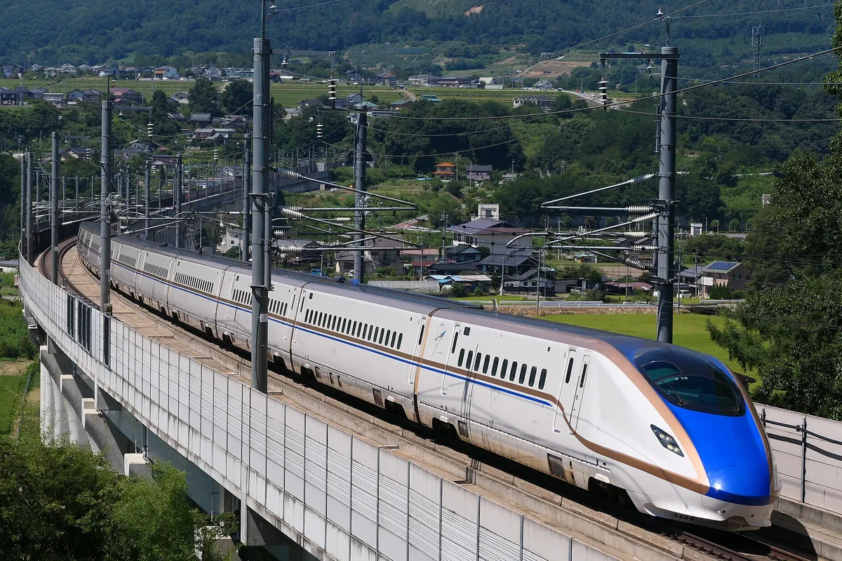 高架橋を走る、青色とベージュの帯が入った白い北陸新幹線の車両が田園風景の中を進む様子の写真