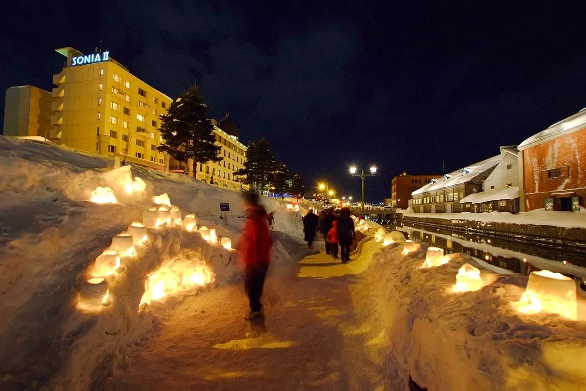 小樽雪あかりの路の運河会場。雪の道に沿って並ぶろうそくの灯りの横を人が歩く夜の写真