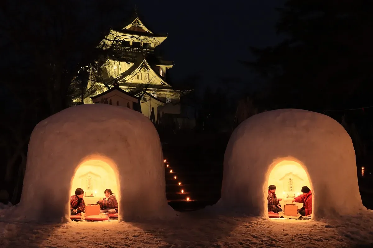 横手のかまくら。横手公園の雪の上に2つのかまくらが並び、中に灯りがともって奥に横手城が見える夕方の写真
