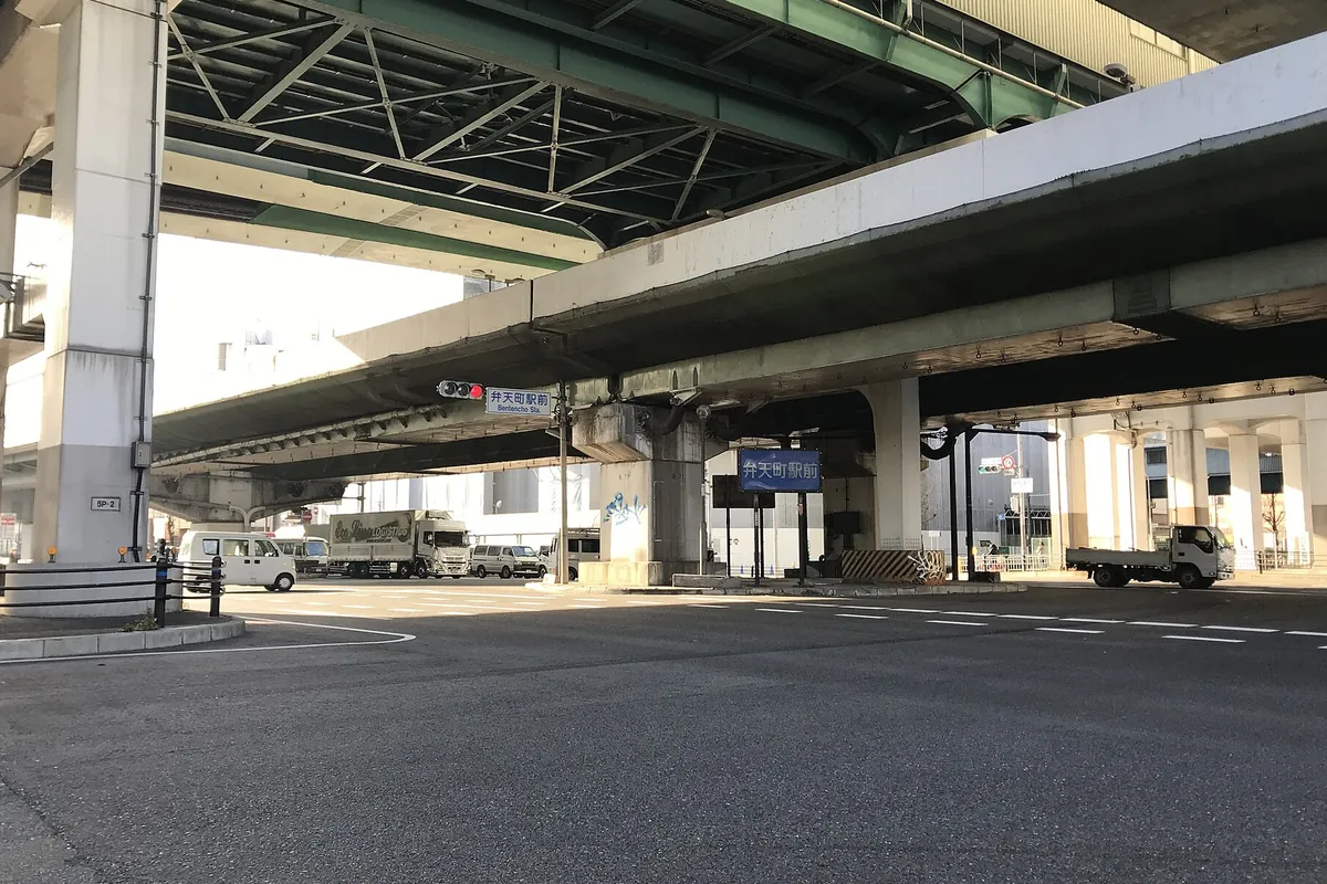 阪神高速の高架道路の下で「弁天町駅前」の案内標識とトラックが写る交差点の写真